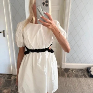 Vit kort klänning med puffärm - Superfin vit kort klänning med puffiga kortärmar och svart volangdetalj i midjan. Klänningen har en luftig passform och är perfekt för varma dagar. Enkel och stilren design som sticker ut med den svarta kontrasten i midjan. Storlek 170 men sitter som S, har lappen kvar