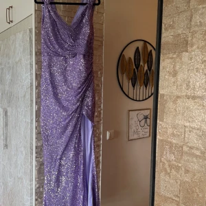 Lila glittrig långklänning med slits - Säljer en lila långklänning med massor av glitter och paljetter. Klänningen har v-ringning, breda axelband och en snygg slits framtill. Figurnära passform som framhäver siluetten och ger en riktigt glam känsla. Perfekt för bal eller festkvällar! Nypris 5000kr använt 1 gång 