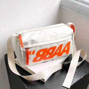 Vit axelväska med orange tryck - Cool vit axelväska med stort orange tryck på framsidan och matchande orange detaljer vid dragkedjorna. Väskan har justerbar rem och praktisk rektangulär form, perfekt för att bära med sig det viktigaste. Tillverkad i slitstark polyester.