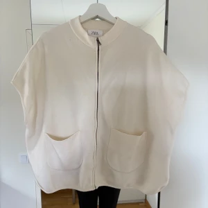Zara poncho  - Stilren beige väst från Zara med rak passform, två stora fickor framtill och dragkedja hela vägen. Västen har mjukt material och breda ärmhål för en avslappnad look. Perfekt att slänga över en hoodie eller t-shirt.