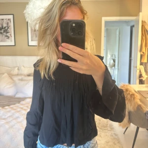 Svart blus med spets och veck - Säljer en svart blus med långa ärmar, fina veck framtill och spetsdetalj vid halsringningen. Blusen har en lös passform och är tillverkad i ett lätt tyg som ger ett snyggt fall. Perfekt att styla med jeans eller kjol för en trendig look.