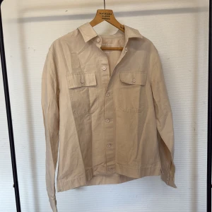 Beige Overshirt från River Island - Stilren beige Overshirt  från River Island Studios i linnemix. Skjortan har klassisk krage, två bröstfickor med lock och knappar framtill. Perfekt för en avslappnad men snygg look. Passar dig som gillar enkel och clean stil.