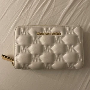 Vit plånbok från Michael Kors - Supersnygg vit plånbok från Michael Kors med quiltad design och präglade MK-loggor. Plånboken har guldfärgad dragkedja och flera fack för kort och sedlar. Perfekt storlek för att ha med sig överallt och ger en lyxig känsla till din vardag.