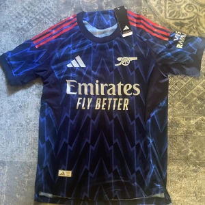 Arsenal blå bortatröja Adidas - Snygg Arsenal bortatröja från Adidas i mörkblått med blixtmönster och röda axelränder. Tröjan har korta ärmar, rund hals och tryckta loggor samt sponsortryck på bröstet. Tillverkad i lätt och ventilerande material, perfekt för match eller träning.