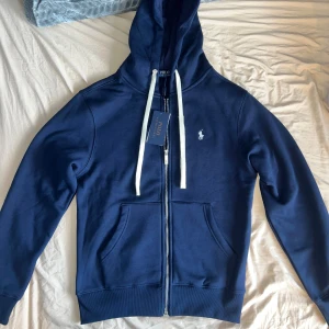 Marinblå Polo Ralph Lauren zipup  - Snygg marinblå hoodie från Polo Ralph Lauren med vit broderad logga på bröstet. Tröjan har dragkedja, huva med vita snören och två stora fickor framtill. Tillverkad i mjuk bomull och perfekt för chill dagar. Klassisk och stilren design. 