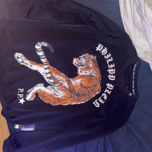 Svart sweatshirt med tiger från Philipp Plein - Cool svart sweatshirt från Philipp Plein med en stor, detaljerad tiger i orange och vitt framtill. Tröjan har lång ärm, rund hals och tryckt logga. Materialet är mjukt och skönt, perfekt för dig som vill sticka ut med en unik design. Storlek L men passar M