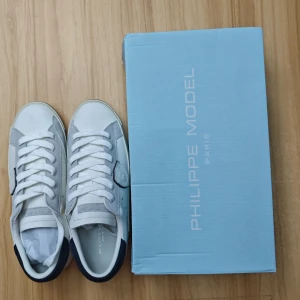 Philippe Model vita sneakers strl 41 - Snygga vita sneakers från Philippe Model med detaljer i grå mocka och mörkblå häl. Klassisk låg modell med snörning och rund tå. Skorna har en platt sula och är tillverkade i skinn och mocka. Perfekta för en stilren och trendig look.