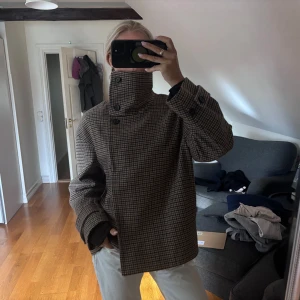 Rutig ulljacka från ARKET - Säljer en snygg rutig jacka från ARKET i bruna och beige toner. Jackan är i ull, har bred krage, dubbelknäppning och stora knappar vid ärmslut. Oversized passform och långärmad, perfekt för höst och vinter. Klassisk och trendig look. Jackan har en defekt som syns på sista bilden men ingenting som syns när man bär jackan!