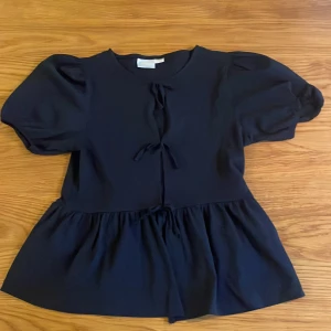 Mörkblå blus med puffärm från Gina Tricot - Supersöt mörkblå blus från Gina Tricot med puffiga korta ärmar och volang nedtill. Blusen har två knytband framtill som ger en extra detalj. Perfekt för dig som gillar en feminin och trendig look.