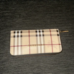 Burberry rutig plånbok med dragkedja - Snygg plånbok från Burberry i klassiskt beige rutigt mönster med svarta, vita och röda detaljer. Plånboken har guldfärgad dragkedja och flera fack för kort och mynt. Perfekt accessoar för dig som gillar stilrena och lyxiga detaljer.