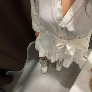 Vit omlottklänning från Zara - Supersöt vit kortklänning från Zara med knappar framtill, volangkant nertill och knytband i midjan. Klänningen har lång ärm och är tillverkad i ett lätt och luftigt material som passar perfekt för varmare dagar.