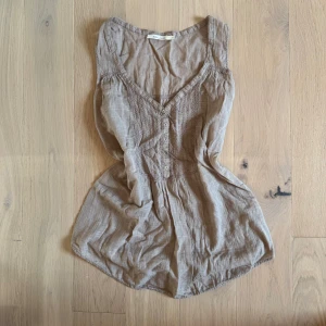 Beige ärmlös blus från Bella Jones - Supersöt beige blus från Bella Jones med v-ringning, knappar framtill och broderade detaljer. Den är ärmlös och har en lätt, luftig känsla med diskret rutmönster i tyget. Perfekt för varma dagar och enkel att styla till jeans eller kjol.