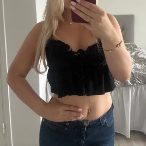 Svart croppad linnetopp med spets - Supersöt svart croppad linnetopp med tunna axelband och spetsdetaljer. Toppen har snörning framtill och volangkant nertill. Perfekt för varma dagar och ger en romantisk vibe till din outfit.