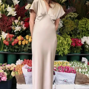 Zara Champagnefärgad långklänning i satin  - Elegant långklänning i champagnefärgad satin med v-ringning och korta ärmar. Klänningen har en mjuk, glansig yta och ett stilrent, flytande fall som ger en lyxig känsla. Perfekt för dig som vill sticka ut med en sofistikerad look.