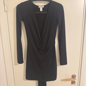Svart kort klänning med djup v-ringning - Snygg svart kort klänning från Cicci & Coco med lång ärm och djup v-ringning framtill. Klänningen har rynkade detaljer vid midjan som ger en cool och smickrande siluett. Perfekt för dig som gillar stilrena och trendiga plagg.