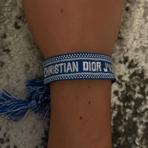 Blått armband från Christian Dior - Snyggt blått armband från Christian Dior med vit broderad text och dekorativa fransar i ändarna. Armbandet har en cool och trendig look som passar perfekt till din outfit. Justerbar storlek med knytning.