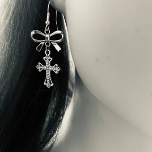 By prisförslag- 1 st Silvrigt örhängen med rosett och kors - Unikat silvrigt örhängen med en söt rosett upptill och ett hängande kors dekorerat med små glittrande stenar. Perfekt accessoar för dig som gillar detaljer och vill sticka ut med en cool och elegant stil.