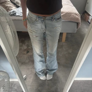 Ljusblå slitna bootcut jeans - Säljer ett par ljusblå jeans med slitningar och bootcut passform. Jeansen har låg midja och klassiska fem fickor. Perfekt för dig som gillar en avslappnad och trendig look. Materialet är jeans och färgen är ljusblå med slitna detaljer.