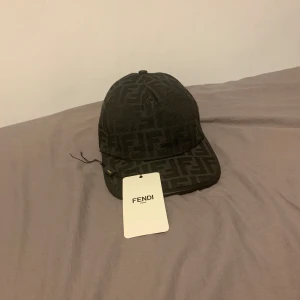 Svart keps från Fendi med logomönster - Snygg svart keps från Fendi med ikoniskt FF-logomönster över hela kepsen. Justerbar baktill med Fendi-detalj. Tillverkad i polyester och bomull för en bekväm och stilren look. Perfekt accessoar för att lyfta din outfit. den är ganska ny och sitter perfekt 