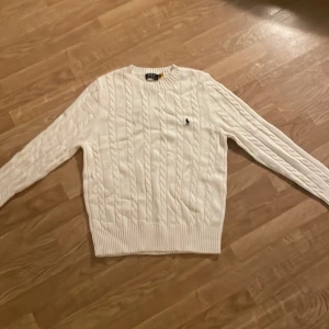 Stickad polotröja - Vit kabelstickad tröja med klassisk rund hals och broderad logga på bröstet. Tröjan har långa ärmar och ribbade muddar vid ärmslut och nederkant. Perfekt för en clean och stilren look.