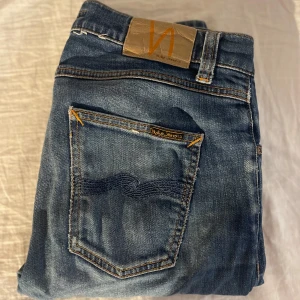 Blåa nudie jeans - Snygga blå jeans från Nudie Jeans med klassisk femficksdesign och orangea sömmar. Jeansen har en rak passform och är tillverkade i slitstarkt denimtyg. Perfekta för dig som gillar en tidlös och avslappnad stil.