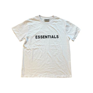 Vit Essentials t-shirt - Vit t-shirt från Essentials med svart tryck på bröstet. Klassisk rund halsringning och korta ärmar. Tillverkad i mjuk bomull för en clean och stilren look. Perfekt för dig som gillar streetwear och enkel design.