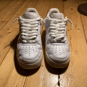 Vita Nike Air Force 1  - Klassiska Nike Air Force 1 sneakers i helvitt skinn med perforerads tå och ikonisk swoosh på sidorna. Lågt skaft, platt sula och snörning framtill. Perfekta för dig som gillar en clean och tidlös look.
