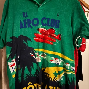 Grön Polo Ralph Lauren Aero Club t-shirt/piké - Exklusiv grön frotté piké från Polo Ralph Lauren med tryck av palmer, flygplan och landskap i färgerna röd, gul, blå och svart. Size: large/XL. Införskaffad 2024 (kvitto finns), använts 2 gånger. Inga synliga skador eller problem. Retail priset när den fortfarande gick att köpa var 2300 kronor (Mitt prisförslag går att förhandlas). Känn dig fri att fråga frågor, jag ska försöka mitt bästa med att besvara dem.