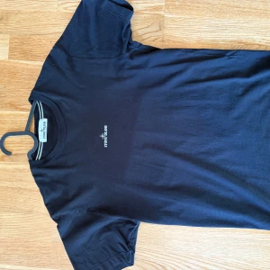 Svart Stone Island t-shirt med tryck - Svart t-shirt från Stone Island i mjuk bomull. Framsidan har en liten vit logga på bröstet och baksidan har ett stort, coolt tryck med Stone Island-text och grafiskt motiv i blått och vitt. Klassisk passform och rund hals.