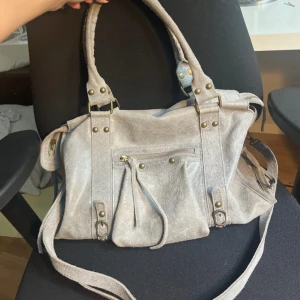 Grå mocka handväska med nitar - Snygg grå axelväska i mocka med mässingsfärgade nitar och detaljer. Väskan har både handtag och lång axelrem, samt en praktisk ficka med dragkedja framtill. 