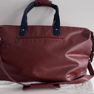 Stor läder weekendbag i skinnimitation - Säljer en rymlig vinröd weekendbag med axelrem och handtag i denimtyg. Väskan har dragkedja upptill, ytterfack med dragkedja och flera innerfack. Perfekt för resor eller gymmet. Materialet är skinnimitation och denim för en cool kontrast.