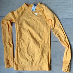 Gymshark träningströja - Figurformad gul långärmad träningströja från Gymshark. Tight passform med tumhål vid ärmslut och  logga på bröstet. Perfekt för gymmet eller löprundan, andas bra och sitter snyggt på kroppen. Helt ny endast testas
