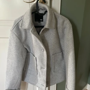 Ljusgrå kappa från 157, XS - Säljer en ljusgrå jacka från 157 i ullblandning. Jackan har klassisk krage, stora fickor framtill och stängs med knappar. Den är lite boxig i modellen och har långa ärmar. Perfekt för dig som gillar stilren och enkel design. Använd fåtal gånger