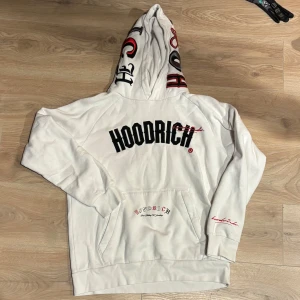 Vit hoodie från Hoodrich  - Vit hoodie från Hoodrich med stor svart logga på bröstet, broderade och tryckta detaljer i rött och svart på huvan och ärmen. Ficka framtill och text på ryggen.