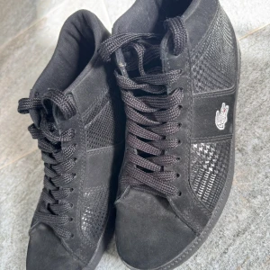 Svarta höga sneakers från Lacoste - Snygga svarta höga sneakers från Lacoste med klassisk krokodillogga på sidan. Skorna har en mix av mocka och syntetmaterial med svagt mönster ton-i-ton på sidorna, rund tå och platt sula. Perfekta för hösten!