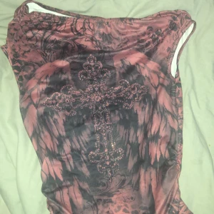 Mörkrosa kortklänning med korsdetalj - Unik kortklänning i mörkrosa och svarta toner med ett stort glittrigt kors framtill. Klänningen har ett coolt mönster med barockinspirerade detaljer och är ärmlös med bred halsringning. Materialet känns stretchigt och följsamt, perfekt för dig som vill sticka ut.