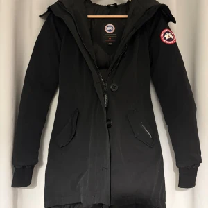 Svart dunjacka från Canada Goose - Ord. Pris 14500kr!! Säljer en svart dunjacka från Canada Goose med klassisk patch på ärmen. Jackan har huva, dragkedja och knappar framtill samt två stora fickor. Insidan är quiltad och fodrad med dun för extra värme. Perfekt för kalla vinterdagar.