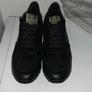 Svarta Valentino Rockrunner sneakers - Säljer ett par helsvarta Valentino Rockrunner sneakers med klassisk design och detaljer i skinn och mocka. Skorna har snörning, rund tå och en platt sula med mönstrad undersida. Perfekta för dig som gillar stilrena och exklusiva sneakers.