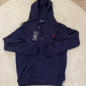 Marinblå hoodie från Polo Ralph Lauren - Snygg marinblå hoodie från Polo Ralph Lauren med dragkedja och huva. Klassisk röd logga broderad på bröstet och ribbade muddar. Tillverkad i mjuk bomull, perfekt för chill dagar. Stilsäker och enkel att matcha med jeans eller joggers. Kan passa som S också.