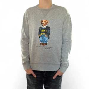 Grå sweatshirt Polo Bear Ralph Lauren - Grå sweatshirt från Ralph Lauren med ikoniska Polo Bear-trycket framtill. Klassisk rund halsringning och långa ärmar. Mjuk bomullskvalitet och normal passform, perfekt för en chill och stilren look.