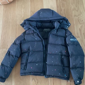 Svart pufferjacka med färgstänk NVLTY - Svart pufferjacka från NVLTY i storlek S med coola färgstänk i vitt, blått och rosa. Jackan har huva, två dragkedjeförsedda fickor och ett stort NVLTY-märke på ärmen. Perfekt för dig som gillar streetstyle och vill sticka ut i vinter.