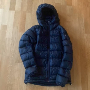 Mörkblå dunjacka Peak Performance XS - Snygg mörkblå dunjacka från Peak Performance i storlek XS. Jackan har huva, dragkedja framtill  en lagning på höger arm