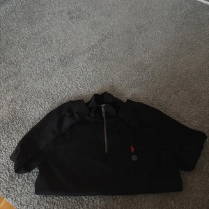 Svart half zip tröja från Ralph Lauren - Svart tröja från Ralph Lauren med half zip och klassisk röd logga broderad på bröstet. Tröjan har hög krage och är långärmad, perfekt för kyligare dagar. Tillverkad i mjuk bomull för en bekväm känsla.