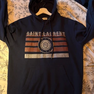 Mörkblå Saint Laurent hoodie med tryck - Snygg mörkblå hoodie från Saint Laurent med stort tryck framtill i rött, vitt och blått. Klassisk känguruficka, huva med snörning och långa ärmar. Perfekt för dig som vill ha en streetig och trendig look. Pris kan diskuteras vid snabb affär! Är en storlek M men sitter som en S