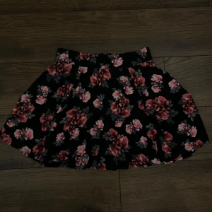 Svart blommig kortkjol med dragkedja - Supersöt svart kortkjol med rosa och röda blommor samt grågröna blad. Kjolen har en markerad midja och en synlig dragkedja bak som ger en cool detalj. Tyget är mjukt och följsamt, perfekt för dig som gillar en feminin och trendig look.