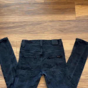 Svarta jeans från Nudie Jeans Co W33 L32 - Svarta jeans från Nudie Jeans Co med klassisk femficksdesign och diskret vågstickning på bakfickorna. Jeansen har svarta sömmar, svart läderpatch i midjan och är tillverkade i mjukt denimtyg. Passar dig som gillar stilrena och bekväma jeans. Jeansen sälj inte längre och är en av dem exklusiva modellerna av tape Ted 