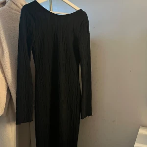 Svart långärmad klänning från SHEIN - Svart långärmad klänning från SHEIN i storlek M. Klänningen har en vågig struktur i tyget, öppen rygg med knyt och är figurnära. Tillverkad i 100% polyester och har en stilren, enkel look med snygga detaljer i ryggen. (Använts max 2 gånger) 