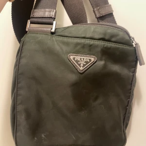 Grön axelväska från Prada i nylon - Snygg mörkgrön axelväska från Prada i slitstark nylon med detaljer i brunt skinn. Väskan har justerbar axelrem, dragkedja upptill och ett lock med metallspänne. Klassisk trekantslogga framtill och innerfack med Prada-märke. Perfekt för dig som vill ha en stilren och praktisk väska.