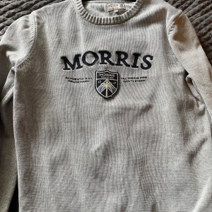 Grå stickad tröja från Morris - Säljer en grå stickad tröja från Morris med broderad logga och emblem på bröstet. Tröjan har rund halsringning och långa ärmar. Perfekt för dig som gillar klassisk och stilren design med en sportig touch.