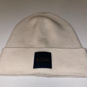 Vit stickad BOSS mössa - Snygg vit stickad mössa från BOSS med uppvikt kant och svart patch framtill. Klassisk och stilren design som passar perfekt nu till vintern. Skriv om ni är intresserade. Mvh Elliott
